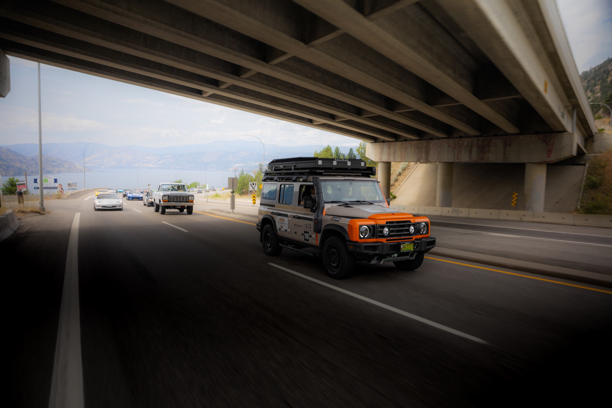 Classic Land Rover Defender driving beneath a bridge with Okanagan Lake beyond